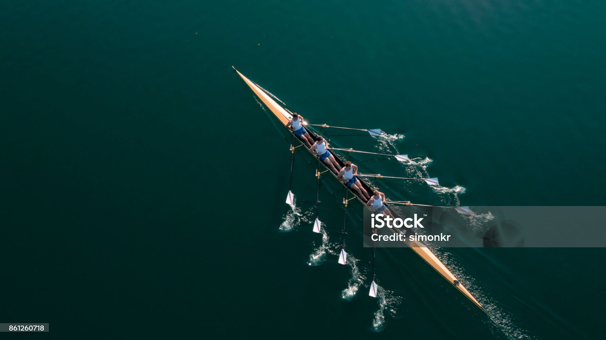 Four male rowers sculling on lake in sunshine.